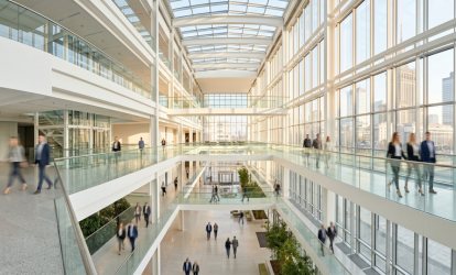 Jasne, wielopoziomowe wnętrze nowoczesnego centrum biurowego z przeszklonymi ciągami komunikacyjnymi, po których spacerują pracownicy. Przez ogromne okna fasady widać architekturę Warszawy.