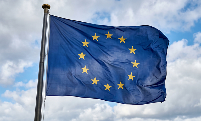 Flaga Unii Europejskiej powiewająca na maszcie na tle błękitnego nieba z białymi chmurami.